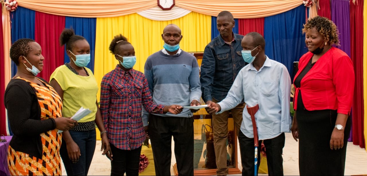 Seven people in a line. Man with crutch receives certificate. He is wearing a shirt and a face mask.